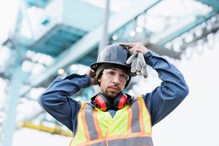 Maritime worker in safety gear adjusting his helmet at an industrial site, representing Longshore vs. Jones Act workers' compensation claims.