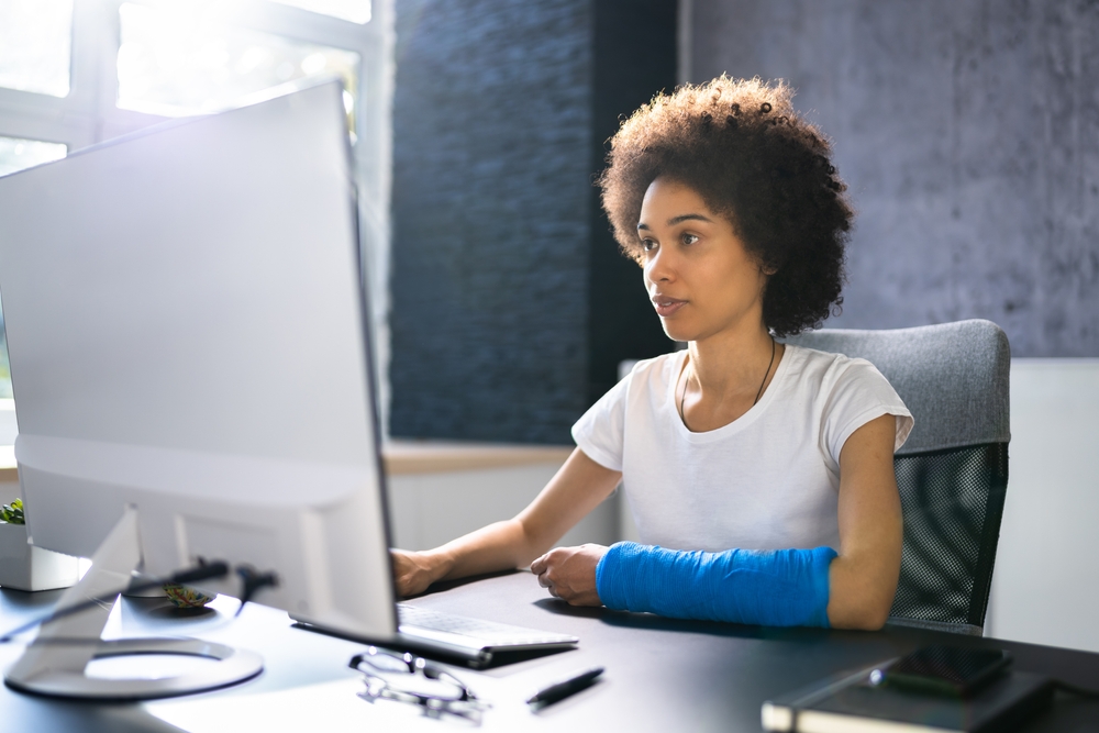 A woman with a blue arm cast working at a desk on a computer, representing an injured employee calculating workers' compensation benefits in Louisiana.