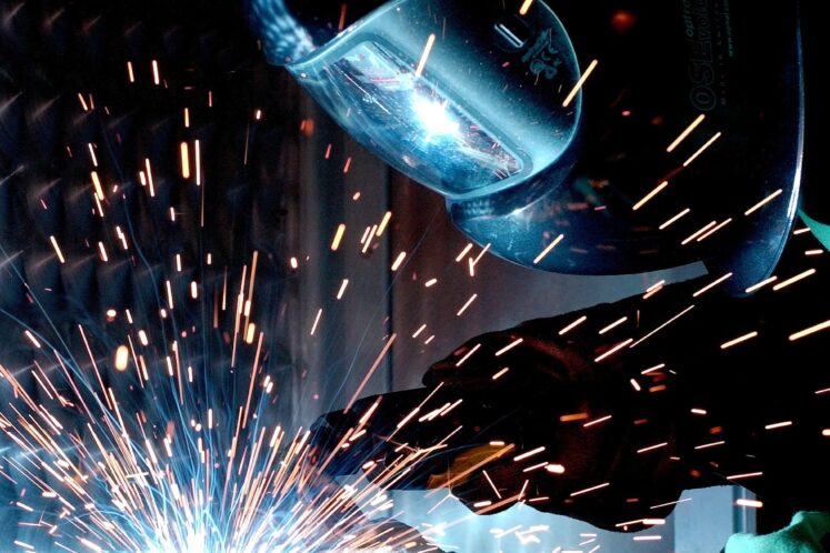 Welder with full gear and mask leans over metal they are welding with sparks flying.